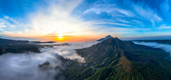 Aerial panorama of Bali’s volcanic highlands at sunrise with a sea of clouds, sun on the horizon, and green ridges in the foreground.