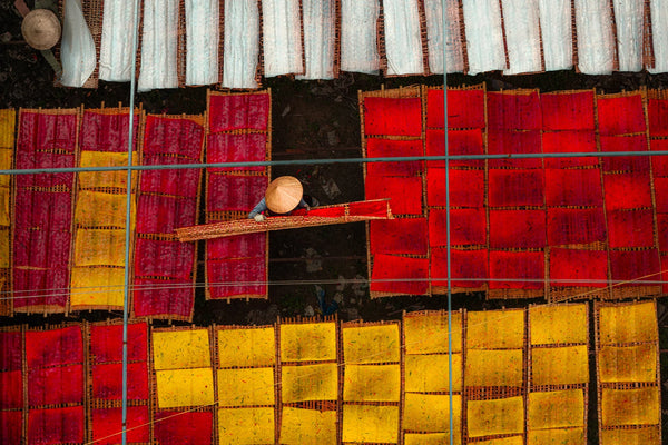 Overhead view of a worker in a conical hat moving a bamboo rack among red, yellow, and white sheets of rice paper and noodles drying in Vietnam.