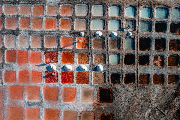 Drone aerial of Omani salt pans with rust-red and aqua basins, workers raking white salt mounds that cast long shadows.