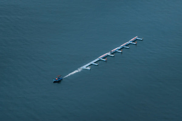 Minimal aerial of Victoria Harbour with a slim blue floating pier of dragon boats and a small motorboat leaving a white wake.