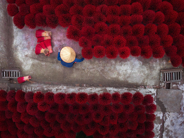 Overhead view of bright red incense bundles drying in rows as a worker in a conical hat moves through a narrow lane.