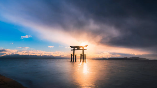 Torii gate of Shirahige Shrine standing in Lake Biwa with the sun shining through and dramatic clouds over the water.