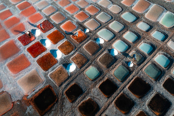 Aerial view of diagonal rows of salt ponds in Oman, punctuated by bright white salt piles and workers casting long shadows.