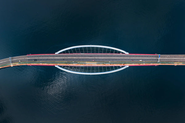 Symmetrical top-down of the Tseung Kwan O–Lam Tin Bridge with a red taxi centered on the roadway over dark blue water.
