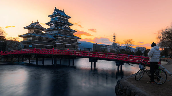 Sunset view of Matsumoto Castle with a red bridge over the moat and a person with a bicycle on the path in the foreground.