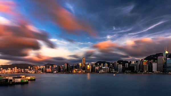 Panoramic view of Hong Kong’s skyline at sunset with fast-moving clouds and warm reflections across Victoria Harbour.