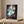 Framed artwork on a wooden table with a pen next to it, against a neutral wall.