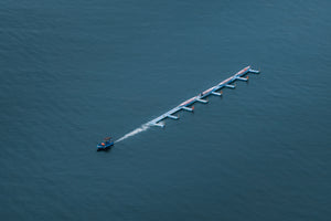 Minimal aerial of Victoria Harbour with a slim blue floating pier of dragon boats and a small motorboat leaving a white wake.