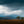 Wide landscape of South African grassland with two small trees on a ridge and dark storm clouds over blue-gray hills.