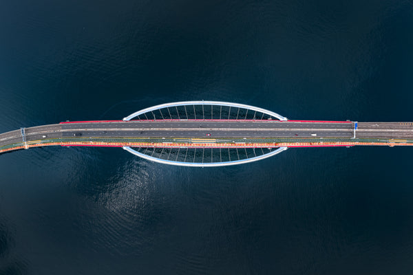 Symmetrical top-down of the Tseung Kwan O–Lam Tin Bridge with a red taxi centered on the roadway over dark blue water.
